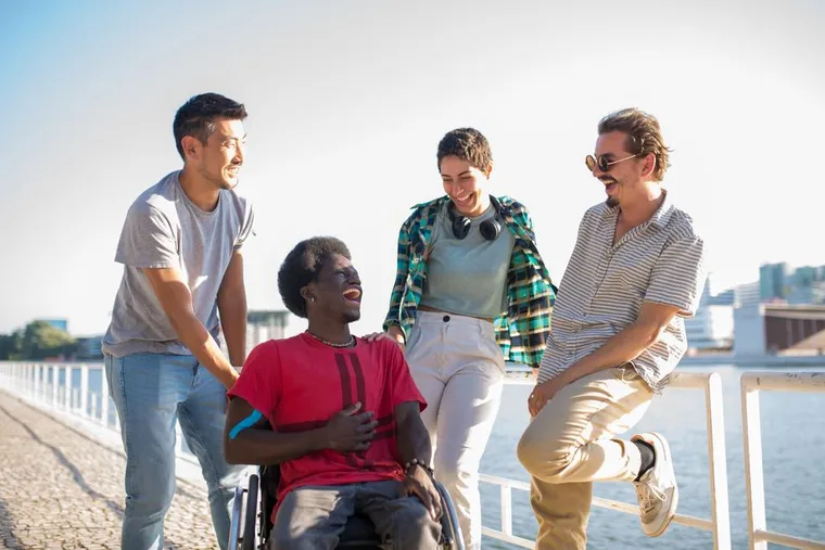 A group of friends laughing together by the water, one using a wheelchair